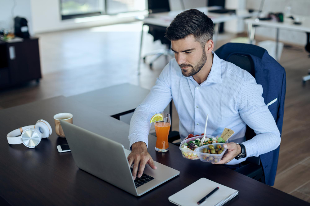 Hombre trabajando frente a su computadora portátil mientras almuerza una ensalada saludable en su escritorio.